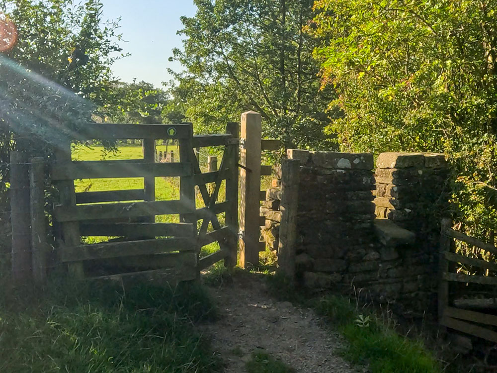 Pass through a wooden kissing gate with a yellow waymarker arrow on it, next to an old stone stile